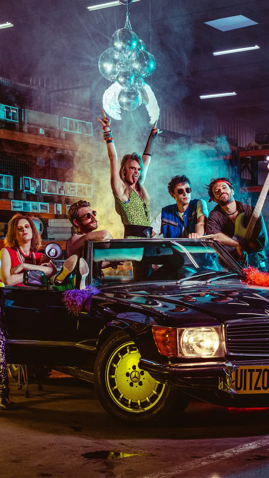 De acht leden van 80's coverband Radio Spandex in kleurrijke jaren 80 outfits rond een klassieke Mercedes cabriolet in een verlichte loods, onder een tros discoballen