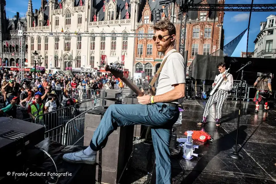 Guitarist Cpt Lee Bido of Radio Spandex plays guitar on stage during Brugge Feest, resting his foot on a monitor speaker with historic Bruges architecture in the background. Photo by Franky Schutz Fotografie.