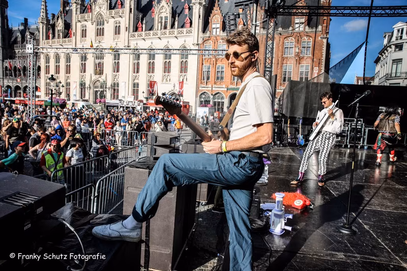 Guitarist Cpt Lee Bido of Radio Spandex plays guitar on stage during Brugge Feest, resting his foot on a monitor speaker with historic Bruges architecture in the background. Photo by Franky Schutz Fotografie.