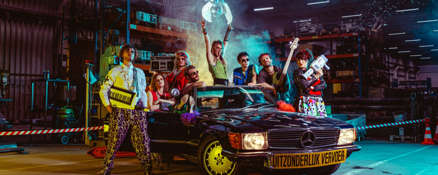 Eight members of 80s cover band Radio Spandex pose in front of a Mercedes oldtimer in a factory warehouse