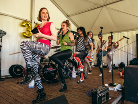Gentle Riverstar en haar koebel trekken het feest op gang — © Michiel Devijver Gentle Riverstar and band members of 80's coverband Radio Spandex performing a choreographed dance with instruments on stage - Michiel Devijver Photography